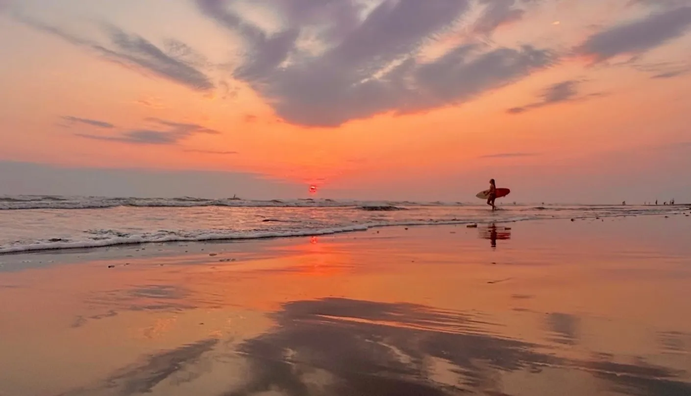 A man holding a surfboard walking on a Costa Rica beach at sunset.