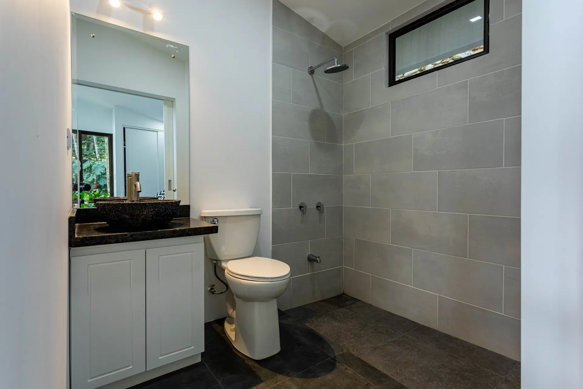 Modern villa bathroom with stone sink, toilet, and walk-in shower featuring large gray tiles and a high privacy window.