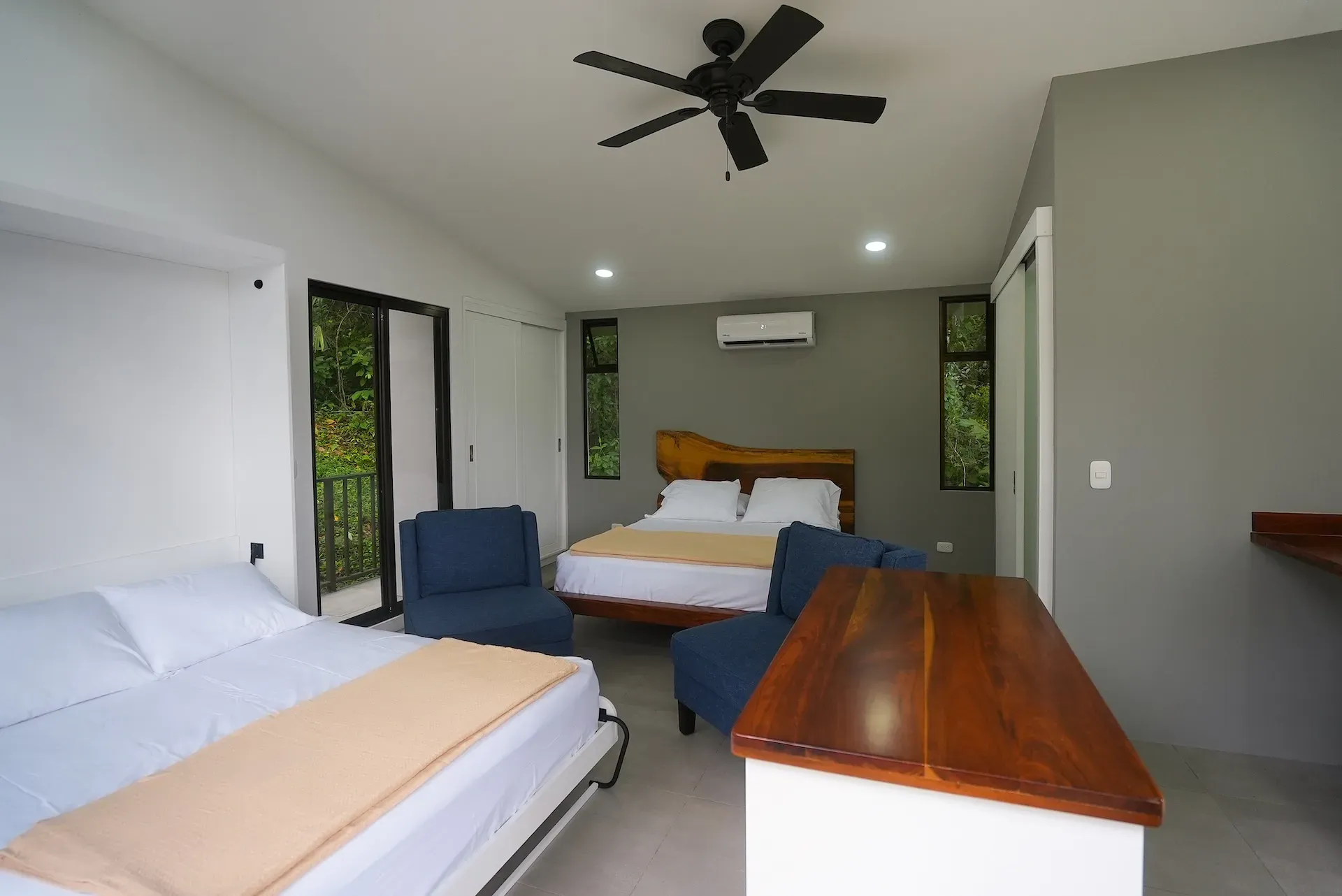 Villa interior with two queen beds, blue chairs, a wooden counter, and sliding doors overlooking jungle greenery.