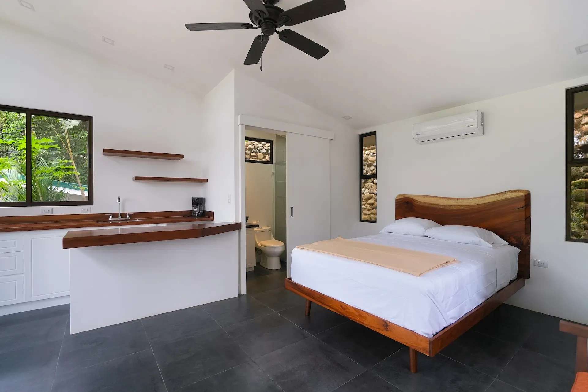 Modern villa room with queen bed, wood-accented kitchenette, ceiling fan, and bathroom entrance.