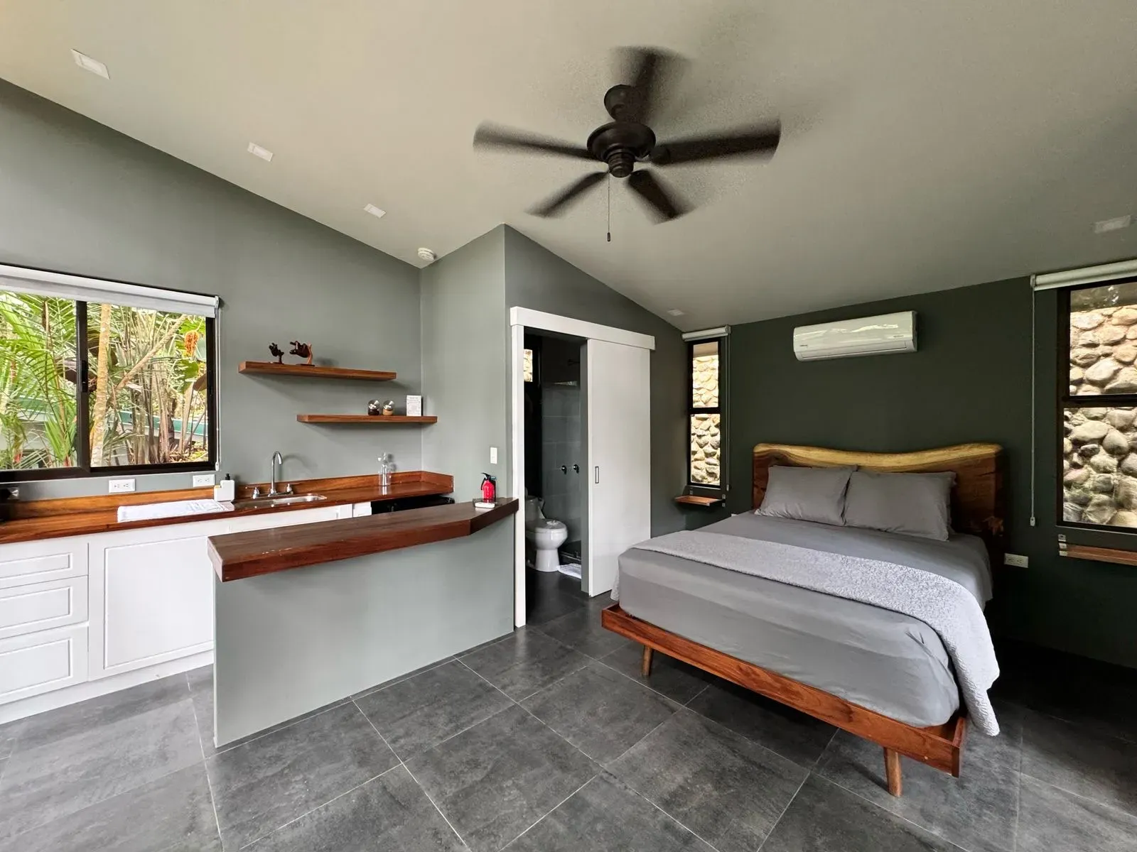 Villa interior with gray bed, wood headboard, kitchenette, and modern dark green walls at Vajra Jahra, Costa Rica.