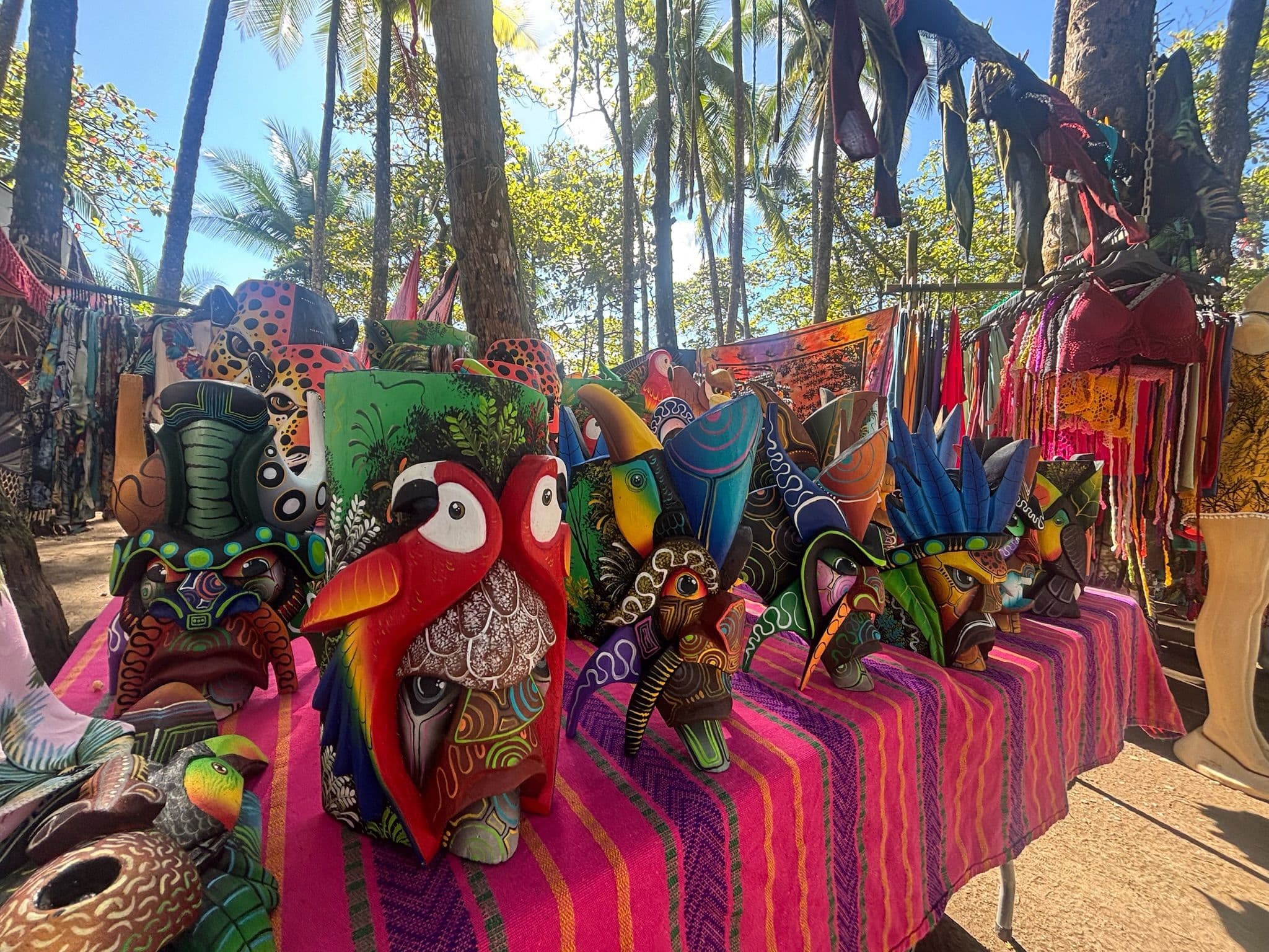 Colorful hand-carved Boruca masks displayed on a vibrant pink textile at an outdoor market in Costa Rica, surrounded by palm trees and local crafts.
