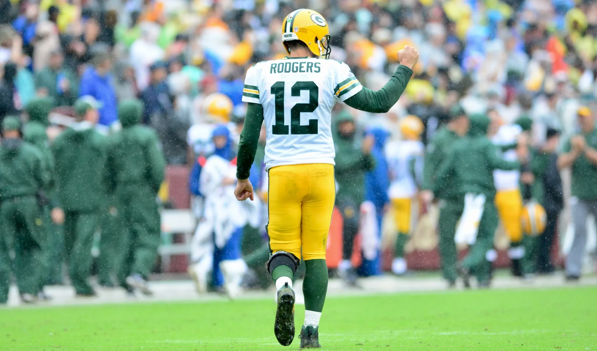 Aaron Rodgers from the back on a football field, in Green Bay Packers uniform, raising his fist with determination.