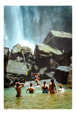 Group of people swimming and exploring near large rocks at the base of a tropical waterfall.