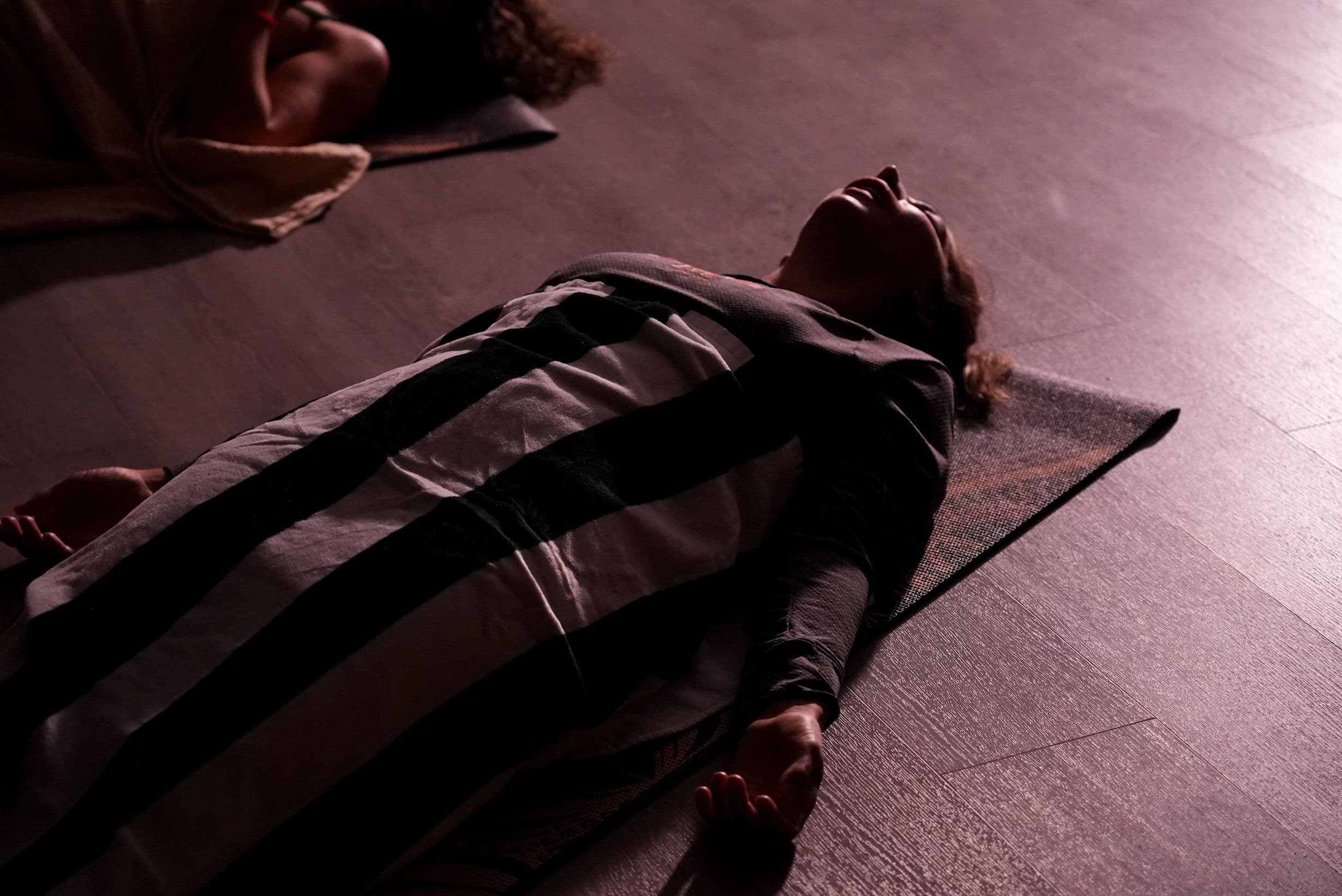 A woman lying on a mat on the floor, in low light, to allow her to relax and focus on healing through sound bathing.