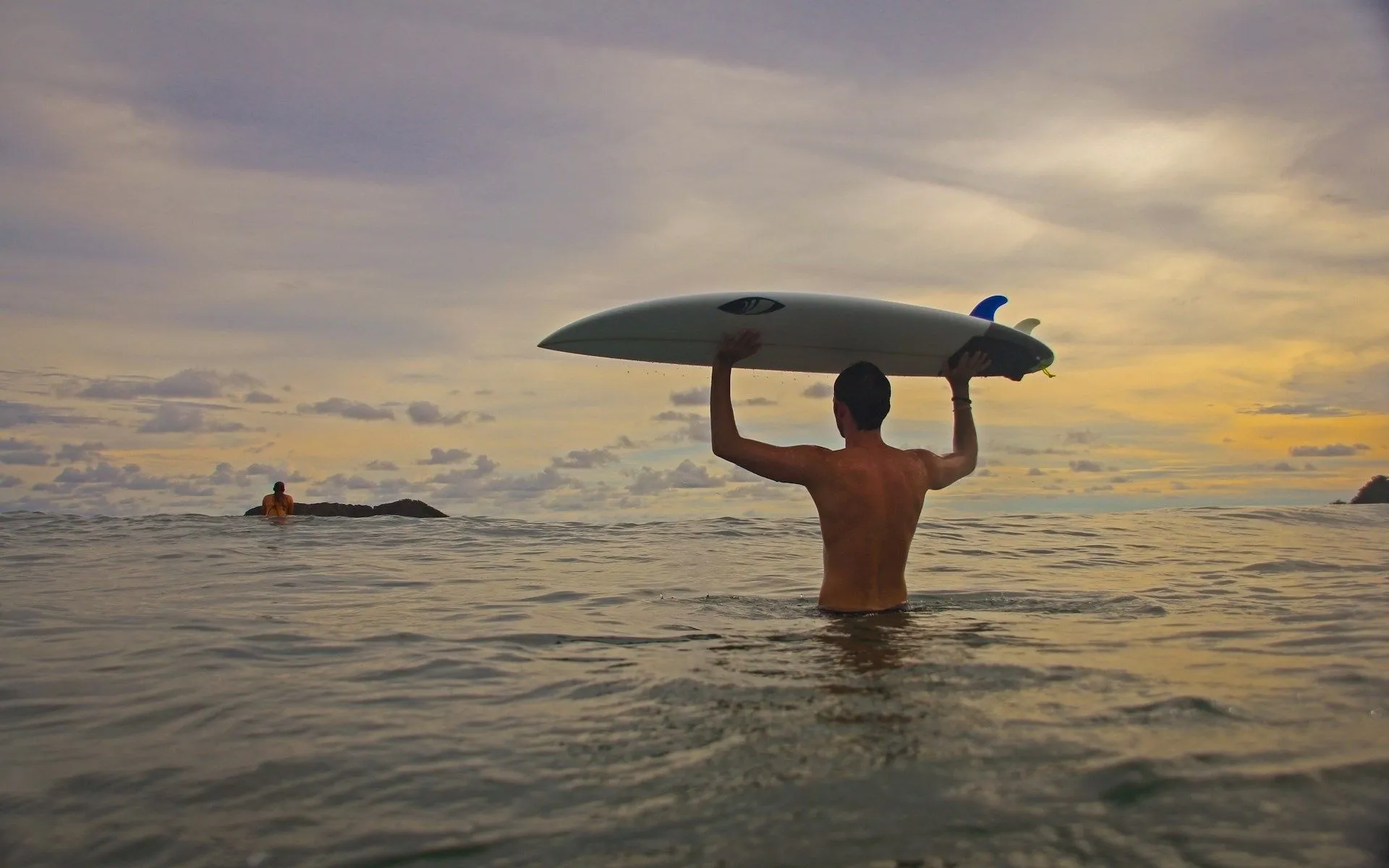 Surfer holding a surfboard in the ocean at sunset near Vajra Jahra Retreat Center, a rejuvenating Corporate Retreat Venue in Costa Rica.