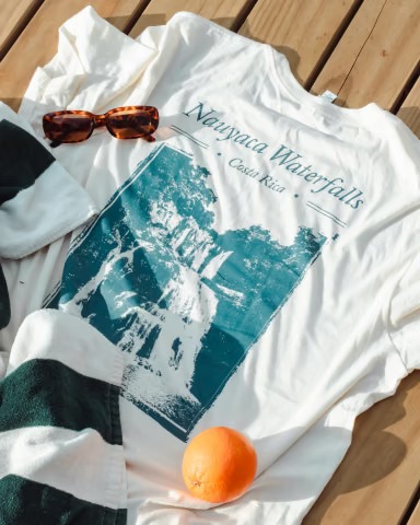 White tee with a Nauyaca Waterfalls, Costa Rica print, laid flat with sunglasses and an orange